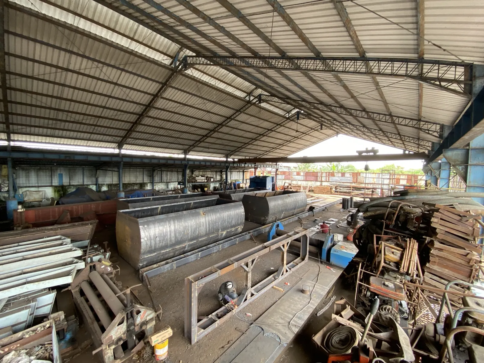 Large steel vessels inside the covered fabrication workshop in Medan, with a welder working on a steel frame in the foreground